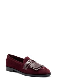 Mocasines de ante burdeos con una correa de cuero marrón contrastante y detalle decorativo de flecos. Suela plana negra. Textura suave, diseño elegante.