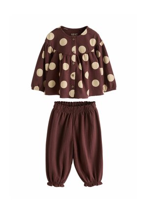 Young child laughing and holding a large cream-colored flower, wearing a white knit hat, brown polka dot top, loose pants, and beige shoes.