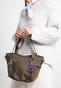 Sac fourre-tout en nylon marron avec des poignées doubles, des poches zippées et un charm en forme de lettre "B" violet. Porté contre un pull blanc texturé.