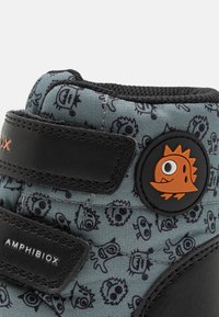 Bottes pour enfants noires et grises avec un motif ludique de visages de dessins animés, accentuées par un logo en caoutchouc et des sangles Velcro.