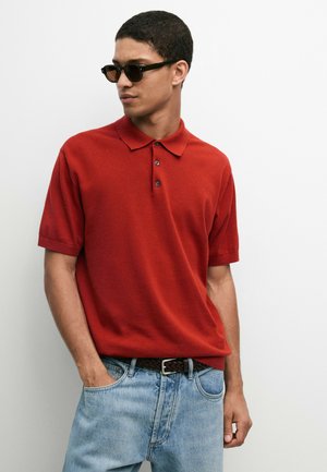 Mann mit schwarzen Sonnenbrillen, rotem kurzärmeligen Poloshirt, hellblauen Jeans und schwarzem Gürtel steht mit einer Hand in der Tasche.
