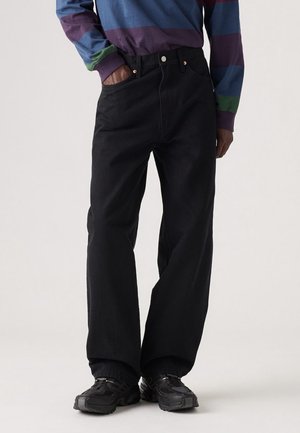 Osoba ubrana w czarne spodnie z szerokimi nogawkami, czarne trampki i koszulę w paski z fioletowymi, niebieskimi, zielonymi i czerwonymi pasami.