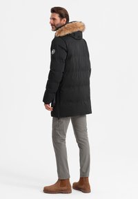 Schwarze Steppjacke mit fellgefütterter Kapuze, steppdesign und Logopatch am Ärmel. Über grauen Hosen und braunen Stiefeletten getragen.