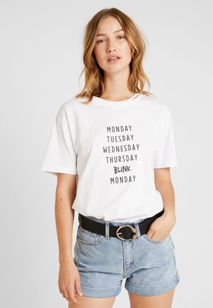 Witte katoenen T-shirt met zwarte tekst die de dagen van de week opsomt, met een ronde hals en korte mouwen, gecombineerd met denim shorts en een zwarte riem.