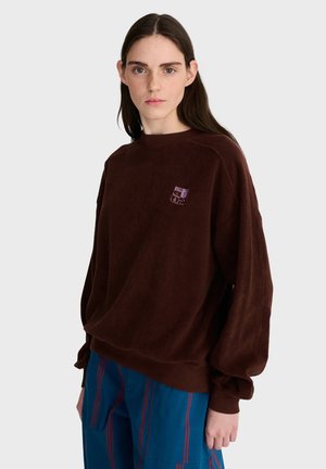 Ung kvinde med langt mørkt hår iført en løs brun sweater med et lille lilla emblem og blå bukser med røde striber.