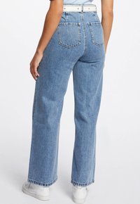 Jeans de mezclilla de un azul claro con una cintura alta, diseño de pierna ancha y bolsillos traseros. El dobladillo presenta un ligero deshilachado, combinados con zapatillas blancas.