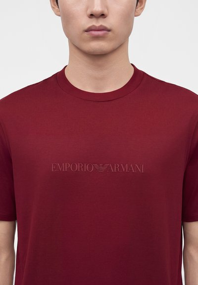 Άνδρας που φοράει απλό μπορντό κοντομάνικο μπλουζάκι με ανάγλυφο λογότυπο Emporio Armani στο στήθος, σε λευκό φόντο.