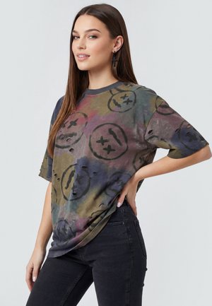 Junge Frau mit langen braunen Haaren trägt ein mehrfarbiges, übergroßes T-Shirt mit abgenutzten Smiley-Prints und schwarze Jeans, posiert mit der Hand in der Hüfte.