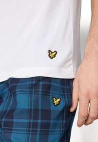 Bela bombažna majica ima črn in zlat izvezeni logotip na robu. Ujemita se z modrimi kratkimi hlačami s karirastim vzorcem, ki prav tako krasijo isti logotip.