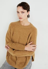 Young person with short dark hair, wearing large hoop earrings, a camel-colored fuzzy sweater, and light grey jeans, arms crossed, neutral expression.