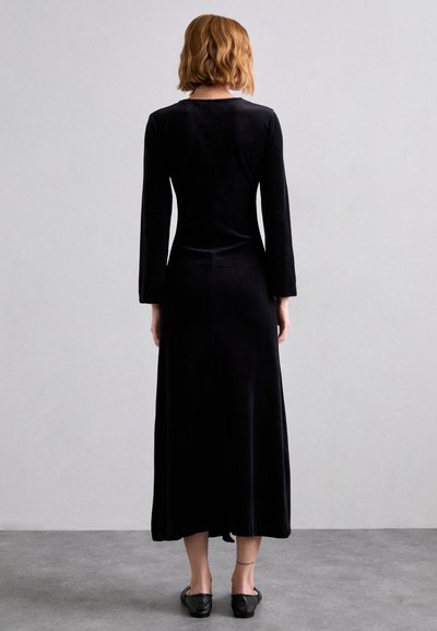 Black velvet long-sleeve dress with a straight cut, reaching mid-calf, featuring a subtle neckline and smooth texture. Worn with flat shoes.