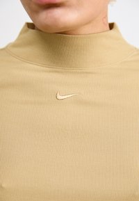 Bežani rebrasti pulover s visokim ovratnikom i suptilnim Nike logotipom izvezenim u sredini. Mekana tekstura s vidljivim vertikalnim linijama.