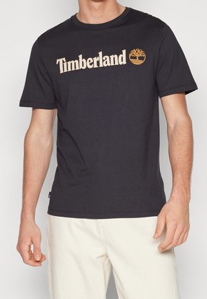 T-shirt Timberland noir à manches courtes avec logo beige et icône d'arbre porté par une personne portant un pantalon clair sur fond blanc.