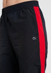 Zwarte sportbroek met een brede rode zijkant. Voorzien van een elastische tailleband en een klein, grijs logo aan de linkerkant. Lichte materialen.