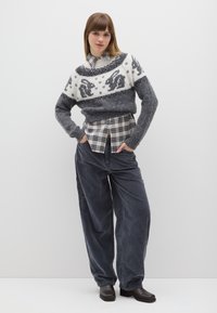 Grauer gestrickter Pullover mit weißem Hasenmuster, darüber getragen ein kariertes Hemd. Weite, graue Cordhosen und dunkle Schuhe mit klobigem Absatz. Schlichter Hintergrund.