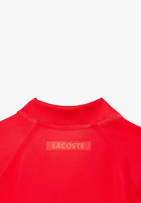 Röd Lacoste-polo skjorta med krage och korta ärmar. Har en mönstrad logotyp på baksidan och ett slätt, lätt tyg.