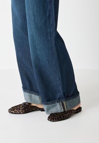 Leopard print mules with a flat sole, featuring a small accent buckle. Paired with cuffed, dark blue, wide-leg denim jeans.