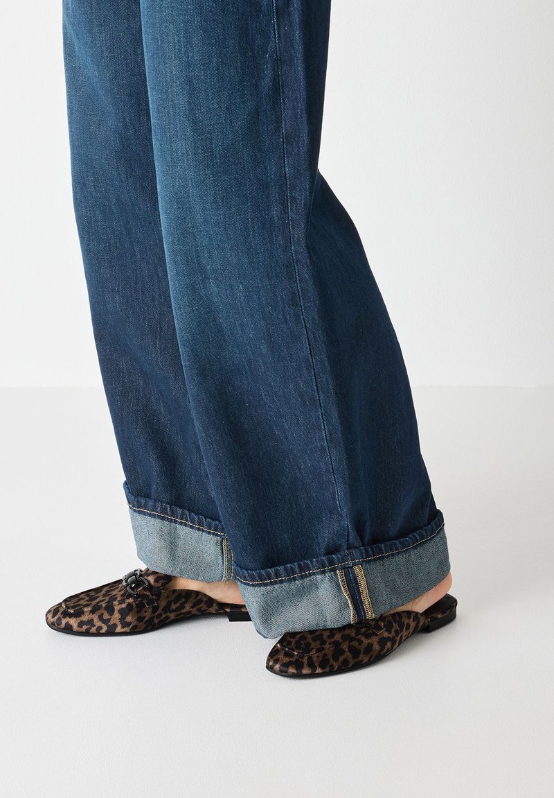 Leopard print mules with a flat sole, featuring a small accent buckle. Paired with cuffed, dark blue, wide-leg denim jeans.