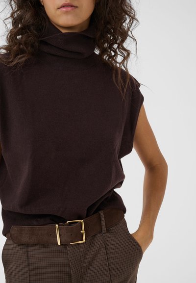 Femme portant un pull sans manches à col roulé marron foncé, un pantalon marron à carreaux et une ceinture marron foncé avec une boucle dorée, main dans la poche.