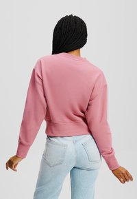 Felpa rosa corta con maniche lunghe, scollo rotondo e polsini a coste, abbinata a jeans di denim azzurri. Texture liscia.