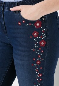 Ciemnoniebieskie dżinsy charakteryzują się misternym haftem kwiatowym w kolorze czerwonym i białym wzdłuż boku, posiadają ukrytą kieszeń oraz gładką fakturę.