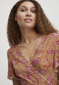 Top avvolgente marrone con motivi floreali rosa e viola, bordi scallop e tessuto testurizzato. Maniche corte e scollatura a V.