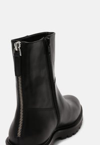 Bottine en cuir noir avec semelle épaisse, dotée de deux fermetures éclair visibles à l'arrière et sur le côté pour la fermeture et le design.