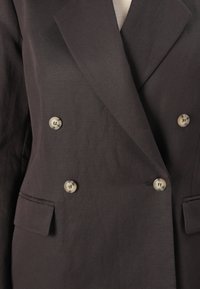 Blazer croisé marron foncé avec quatre boutons beiges et deux poches à rabats, porté sur un top côtelé beige clair.