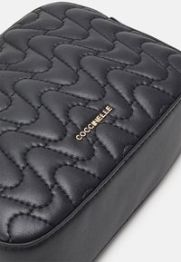 Czarna kosmetyczka ze skórki pikowanej o gładkiej fakturze, z złotym logo "Coccinelle" i zaokrąglonymi krawędziami.
