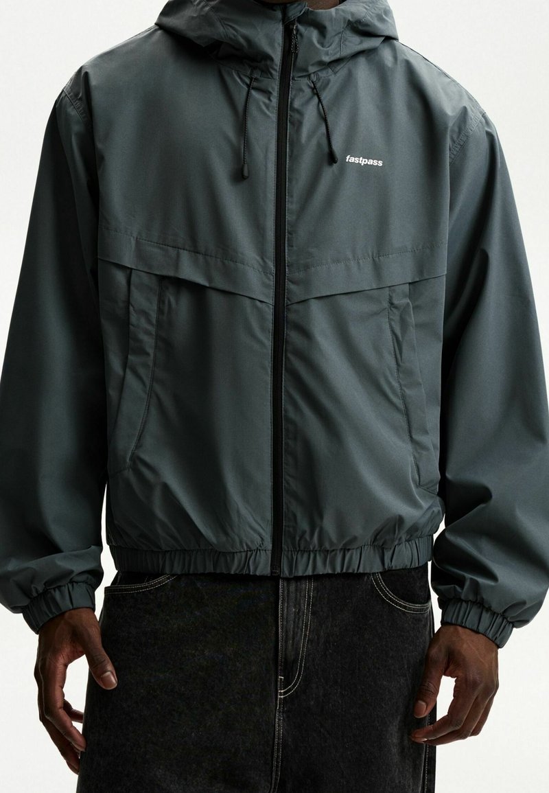 Man wearing dark gray hooded zip jacket with "fastpass" logo on chest and elastic cuffs, paired with black pants.