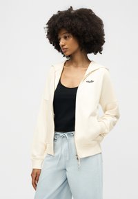 Γυναίκα με σγουρά μαλλιά που φοράει κρεμ φούτερ με φερμουάρ πάνω από μαύρο τοπ και ανοιχτό μπλε παντελόνι με κορδόνι, στέκεται μπροστά σε απλό φόντο.