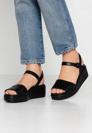 Fødder iført sorte plateau-sandaler med ankelremme sammen med blå korte jeans, stående på en hvid overflade.
