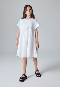 Hvid bomuldskjole med korte ærmer, rynket overdelsstykke og en kant med bølget blonder. Bæres med sorte sandaler med stropper.