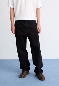 Pantalones de algodón negros de corte suelto, con cintura alta y bolsillos laterales, combinados con zapatos de cordones marrón oscuro. Camiseta blanca sencilla arriba.