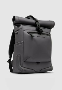 Mochila gris con cierre enrollable, fabricada con material resistente y repelente al agua. Cuenta con correas ajustables, un bolsillo frontal con cremallera y detalles texturizados únicos.