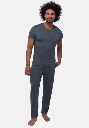 Lachende man met krullend haar, gekleed in een aansluitend donkergrijs T-shirt met korte mouwen en bijpassende broek, staat blootsvoets tegen een effen achtergrond.