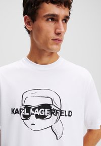 Biela bavlnená tričko s čiernym grafickým motívom postavy v slnečných okuliaroch a nápisom "KARL LAGERFELD." Strih s okrúhlym výstrihom, krátke rukávy.