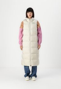 Lange puffer gilet in licht beige met een doorgestikt ontwerp, hoge kraag en ritsluiting. Draagt over een roze en bruine langarmtop en een spijkerbroek.