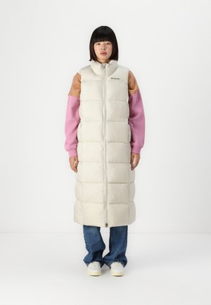 Lange puffer gilet in licht beige met een doorgestikt ontwerp, hoge kraag en ritsluiting. Draagt over een roze en bruine langarmtop en een spijkerbroek.