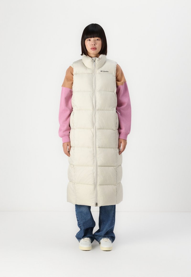 Lange puffer gilet in licht beige met een doorgestikt ontwerp, hoge kraag en ritsluiting. Draagt over een roze en bruine langarmtop en een spijkerbroek.