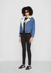 Veste en denim avec col en fausse fourrure, couleur bleue, fermeture éclair à l'avant, portée sur un t-shirt graphique noir, associée à des leggings noirs et des baskets.