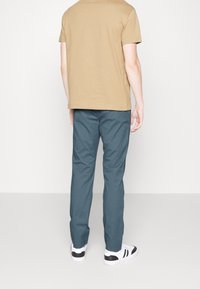 Beige t-shirt med korta ärmar, kombinerad med teal chinos och svart-vita sneakers, sedd bakifrån. En enkel och avslappnad design.