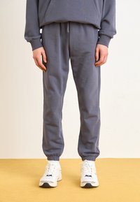 Grå joggebukser laget av bomull, med elastisk midje og mansjetter, sammen med hvite joggesko med blå detaljer.