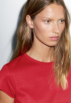 Jonge vrouw met steil blond haar en groene ogen, gekleed in een felrood shirt met korte mouwen, kijkt iets naar rechts met een neutrale uitdrukking.