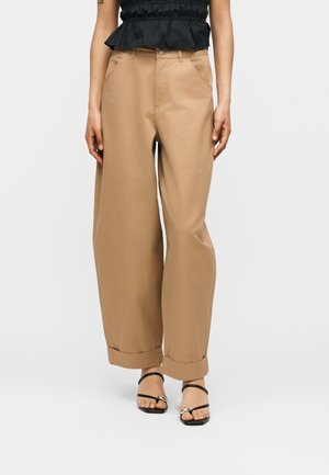 Femme portant un pantalon beige taille haute à jambes larges avec des revers roulés, un haut noir sans manches et des sandales noires à brides, debout sur fond blanc.