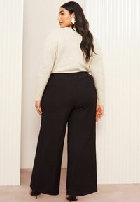 Plus-size model nosi svijetlu bež, mekano teksturiranu džempericu i crne hlače širokih nogavica, pokazujući opušteni kroj i nabran pojas.