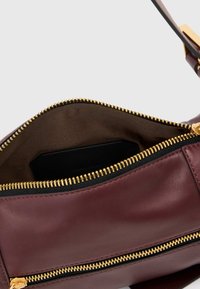 Bordeauxrode lederen tas met een gouden ritssluiting, voorzien van een ruim met stof gevoerd interieur en een kleine zwarte kaarthouder die zichtbaar is aan de binnenkant.