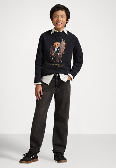 Granatowa bluza z kapturem z grafiką misia w stroju formalnym, zestawiona z czarnymi dżinsami i czarnymi trampkami z białymi akcentami.