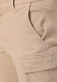 Shorts cargo beiges en tissu mélangé coton, comportant une poche latérale, des poches avant inclinées et des détails de couture visibles.