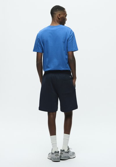 T-shirt en coton bleu associé à un short bleu marine foncé. Présente un design simple, une coupe décontractée, avec des poches latérales et une taille côtelée. Baskets et chaussettes visibles.